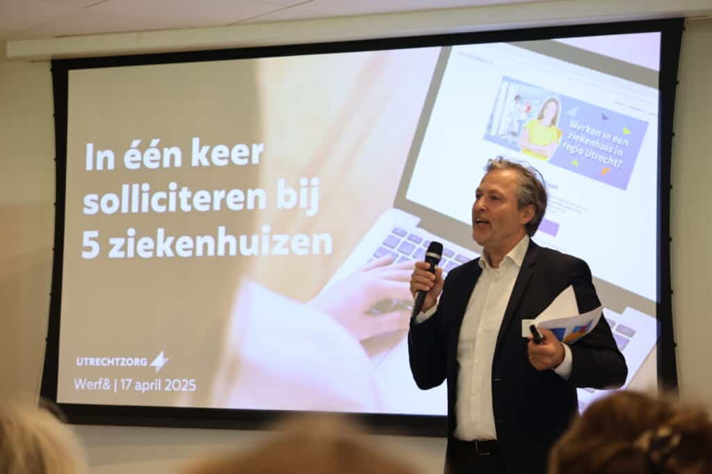 Een man staat voor een groot scherm. Hij houdt een microfoon in zijn handt en is iets aan het vertellen. Hij heeft een pak aan. Op het scherm staat "In één keer solliciteren bij 5 ziekenhuizen".