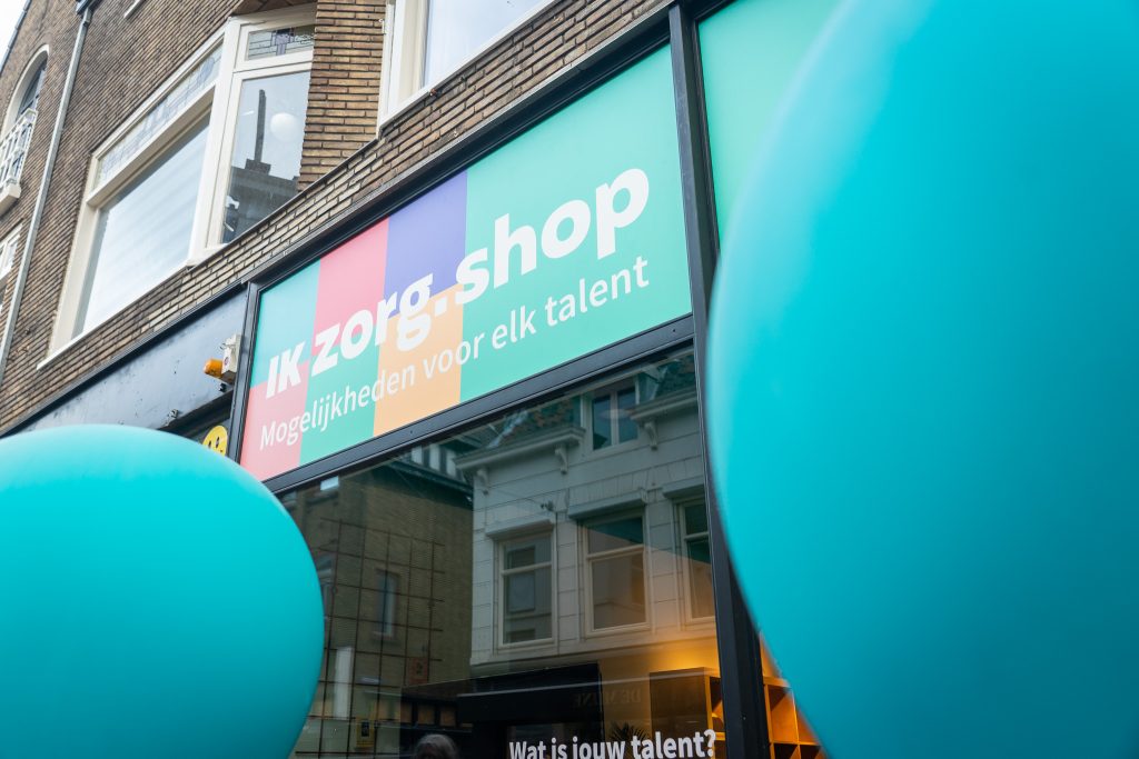 Je ziet de voorgevel van de Ik Zorg Shop in Utrecht: een bord met verschillende gekleurde blokken waar met witte letters 'Ik Zorg.Shop - Mogelijkheden voor elk talent' op staat. Aan de zijkanten zie je lichtblauwe ballonnen.
