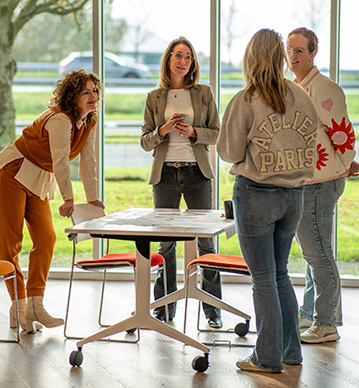 Vier vrouwen staan om een witte tafel heen in een lichte ruimte met grote ramen op de achtergrond.