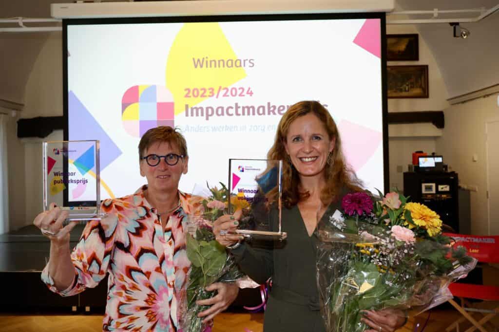 Twee vrouwen staan voor een scherm met de titel Winnaars 2023/2024 Impactmakers. Ze hebben een oorkonde vast en bloemen.